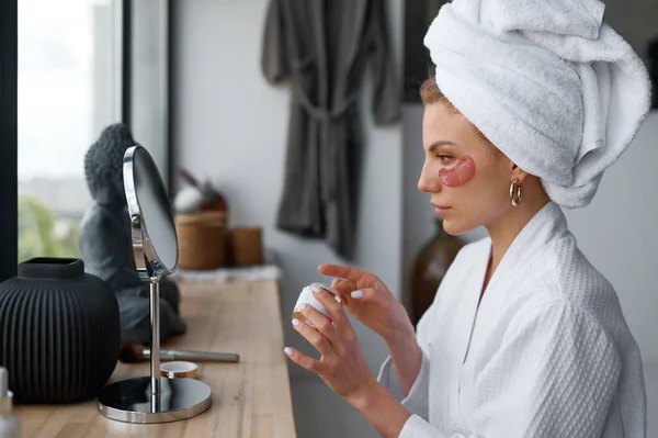 Young woman in towel applying cosmetic beauty patch under eyes to remove wrinkles after taking bath or shower. Skin care, beauty treatment, cosmetology, dermatology