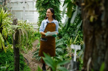 Young female gardener working in greenhouse growing tropical plants. Woman making notes in notebook during research experiment