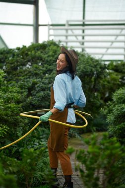 Happy young woman gardener having fun watering plants with hose in greenhouse. Farming, gardening, agriculture concept