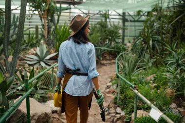 Young woman gardener in overalls leaving greenhouse after work. Attractive female florist walking along footpath view from back