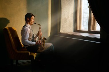 Jazz musician playing saxophone at the window sitting in chair in retro room
