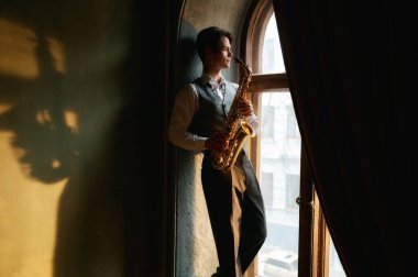 Young man with saxophone standing on windowsill of old room in daylight.