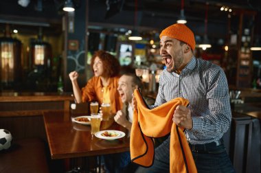 Two guys and woman at pub watching football game and drinking beer. Young excited overjoyed people shouting feeling crazy. Focus on mad man gesticulating with scarf in hands and screaming