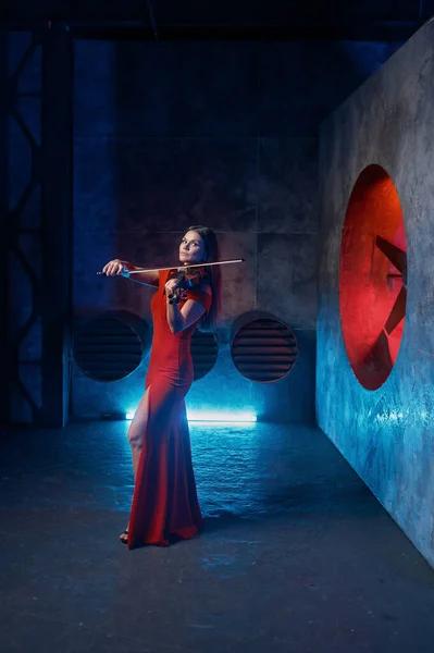 Female violinist performing on grange stage for camera. Woman musician performer wearing elegant red dress playing classical music against industrial background