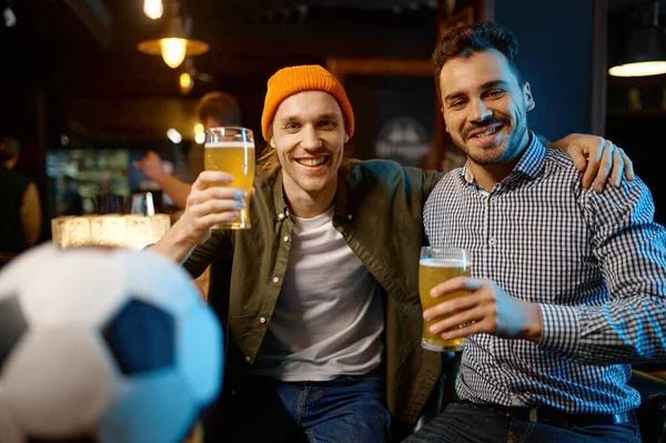 Portrait of friends football fans embracing and looking at camera. Young people drinking beer while watching soccer match championship in sports bar