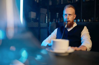 Handsome bearded man sitting at table and smoking hookah while drinking coffee in bar. Selective focus