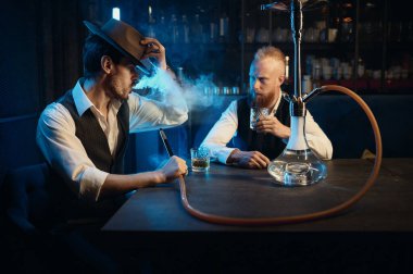 Retro well-dressed gangsters smoking hookah in bar. Serious handsome guys having business meeting while spending time at nightclub
