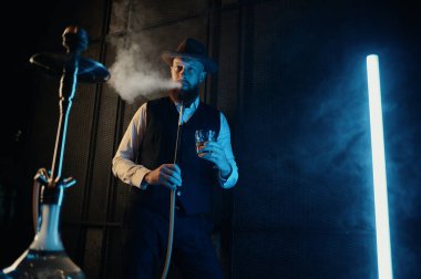 Front view of resting man smoking hookah at home workplace. Retro guy in hat drinking relax in dark room with neon lighting