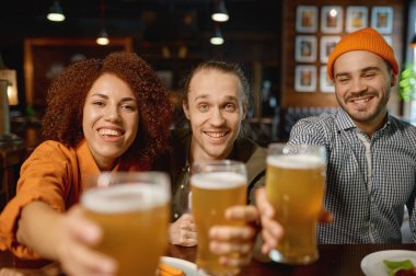 Group of young people smiling at camera and clinking glasses. Happy multiracial friends toasting with beer. Meeting in sports bar