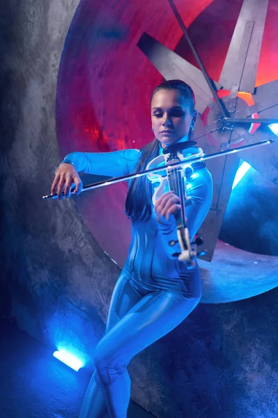Young woman playing magic melody on electronic violin in neon glowing light standing against industrial air blower turbine