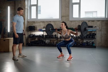 Young sportswoman training bicep with heavy barbell under control of professional fitness instructor
