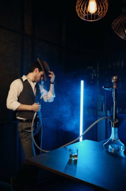 Retro dressed guy with hat smoking hookah while standing in clouds of smoke and looking at camera