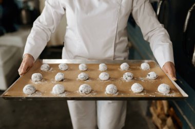 Üniformalı fırıncı hamurdan yapılmış kurabiyelerle tepsi taşıyor ve pişirmeye hazırlanıyor. Hamur işi üretimi