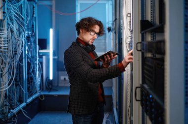 Dijital tabletli genç erkek bilişim uzmanı bilgisayar sunucu yapılandırmasını kontrol ediyor. Modern veri merkezi
