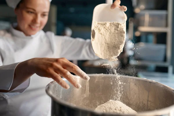 白い制服を着たプロの女性のお菓子は お菓子のテーブルの近くに立って大きな金属製のボウルに小麦粉を入れて クローズアップビュー 選択フォーカス — ストック写真