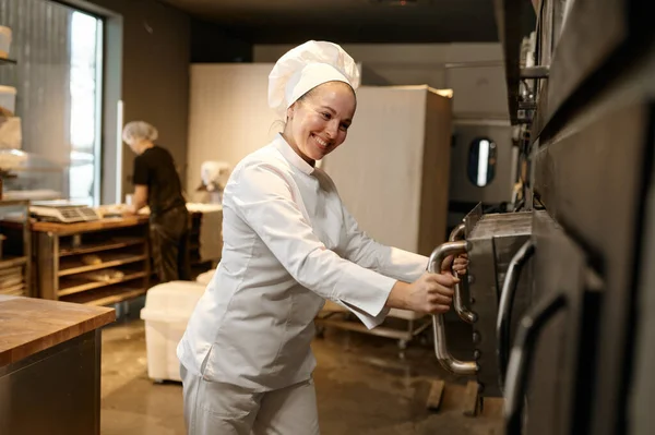 現代のパン屋キッチンで大きな産業オーブンを開く制服を着た若い笑顔の女性パン屋 ベーカリーハウスでのパンとペストリーの生産 — ストック写真