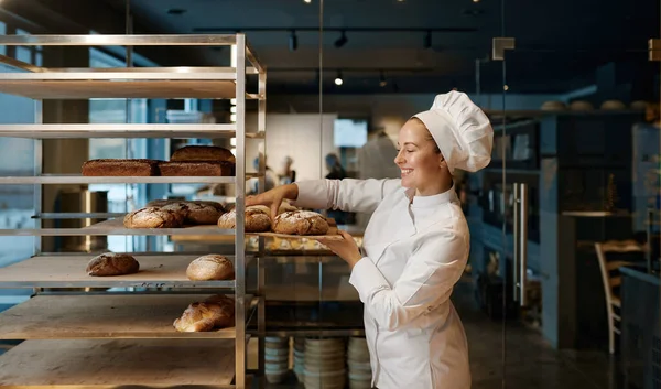 Beautiful woman baker in uniform checking freshly baked bread on tray rack at modern bakery kitchen
