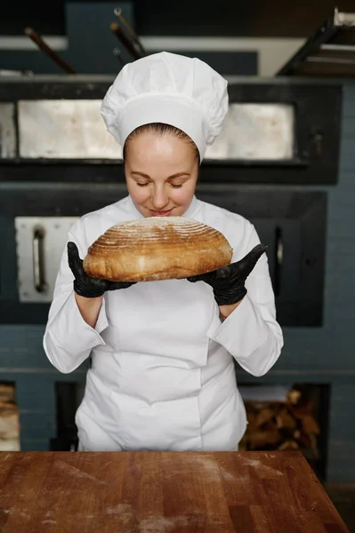 若い女性のパン職人は 現代の商業ベーカリーハウスの背景に新鮮なパンを嗅いでいます 焼き菓子の生産 — ストック写真