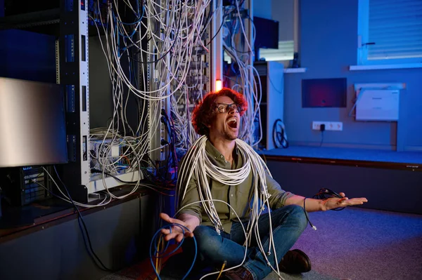Stressed computer engineer yelling while looking up sitting wrapped in optic cables trying to understand network mess