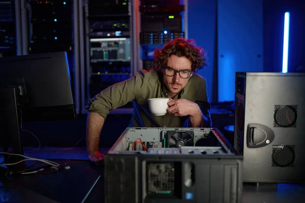 分解されたコンピュータシステムユニットを見てコーヒーを飲む修理工 サーバールームの問題を解決する集中的過労の男性技術者 — ストック写真