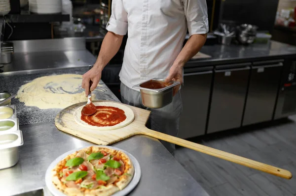 ピザベースにトマトペーストを広げる男性シェフ おいしいレシピで伝統的なイタリア料理のベーキングプロセス — ストック写真
