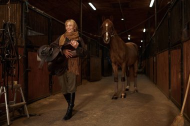Female horse rider walking with harness in stable. Young adult woman rancher going with saddle in hands and smiling
