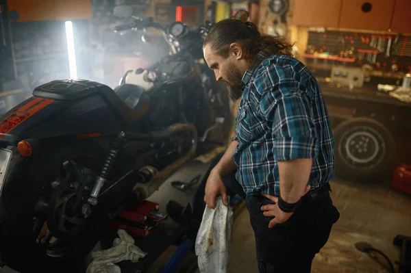 修理工場でオートバイのエンジンを修理している間 彼の手を拭く残酷なバイカー バイクのメンテナンスコンセプトに取り組む髭の男の整備士 — ストック写真