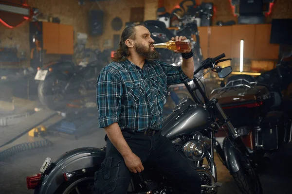 ガレージでのハード修理作業の後 ボトルからビールを飲む疲れた男バイカー 大人の男の肖像画彼のオートバイに座って — ストック写真