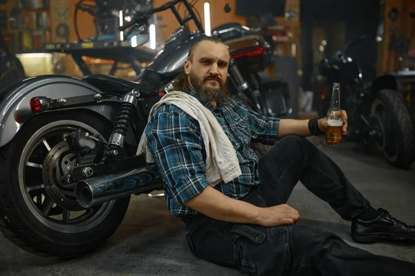 Exhausted mature man biker sitting on floor holding opened glass of beer after hard long work in garage. View from above
