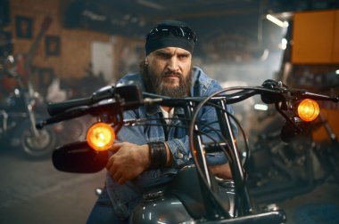 Garaj mağazasında yeni motosiklet deneyen yakışıklı sakallı adam. Motor atölyesinde yeni bir araç seçen motorcu