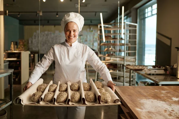 現代のパン屋のプロキッチンで働きながら ゴマ入りの甘いパン菓子を準備する笑顔の女性パン屋 — ストック写真