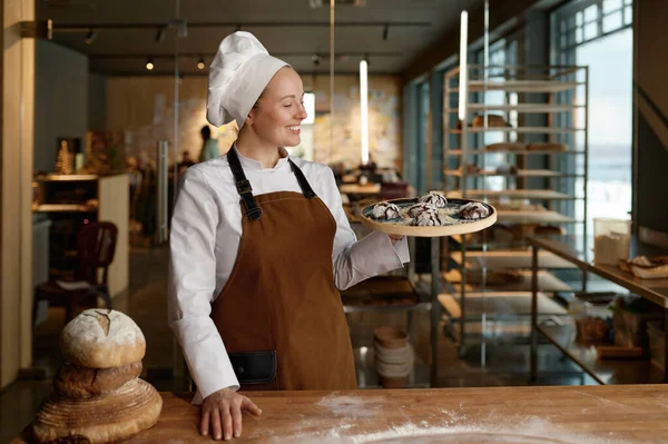 Young female pastry chef presenting freshly baked cookies standing at bakery kitchen. Happy confectioner showing delicious freshly made sweet brownie on tray