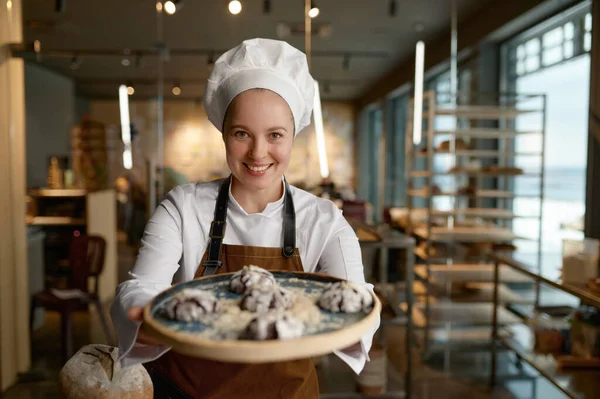 Smiling female pastry chef presenting freshly baked cookies standing at bakery kitchen. Happy confectioner showing delicious freshly made sweet brownie on tray