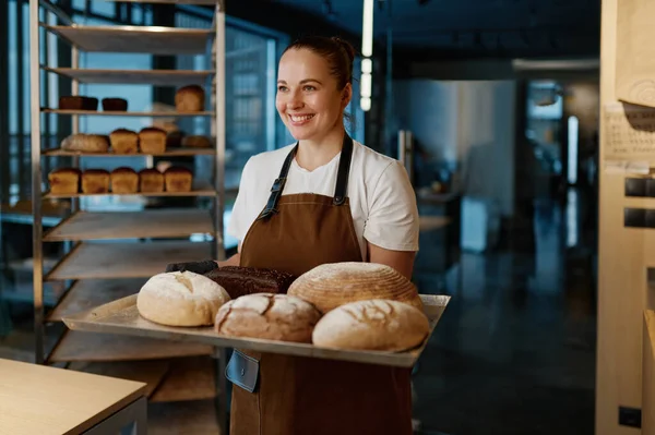 Pretty woman baker holding variety of baked bread on tray standing over bakery house background