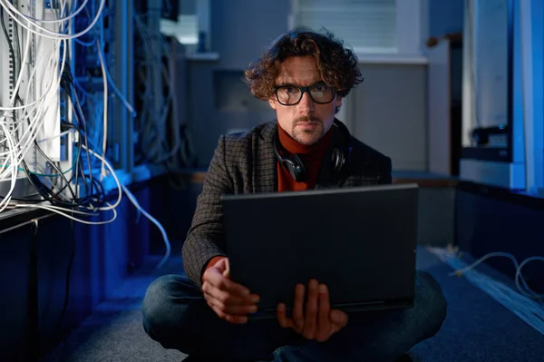 Portrait of male system administrator working in server room using laptop while inspecting in computer. Database management and maintenance concept