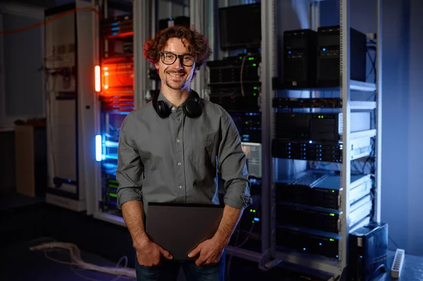 Portrait of young smiling network engineer standing with laptop in server room and looking at camera