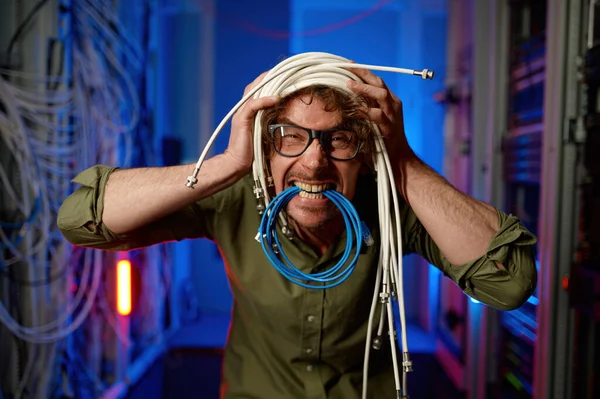 Funny crazy system administrator with head wrapped in wires and optical cables in teeth fooling around in server room. Comic face portrait of overworked computer engineer