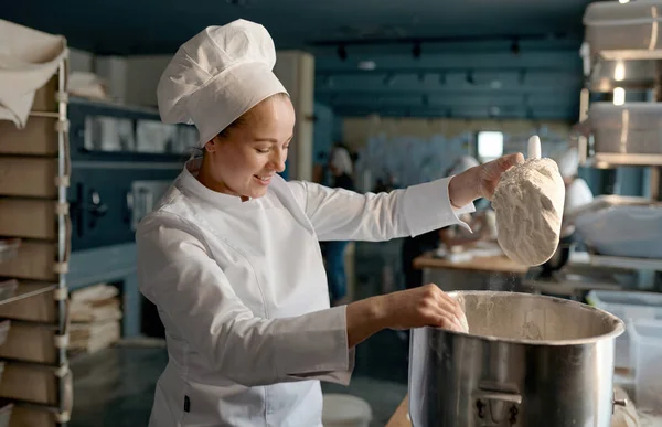 お菓子のテーブルの近くに立つ大きな金属製のボウルに小麦粉を入れて白い制服を着たプロの女性のお菓子 — ストック写真