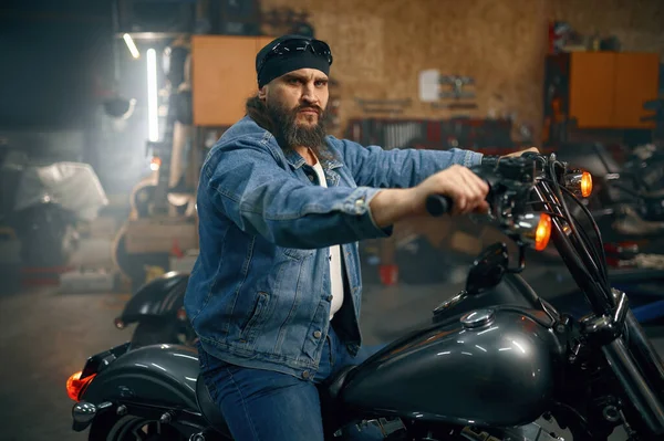 ガレージのインテリア上のオートバイに成熟した髭バイカーの肖像画 真剣に残忍な剃毛男バイクライダー見ますカメラ — ストック写真