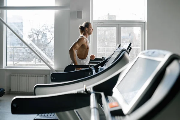 現代のスポーツジムでトレッドミルを走る強い筋肉質の男 カーディオトレーニングを行う固定機械上のアスレチック男性ボディービルダージョギング サイドビュー — ストック写真