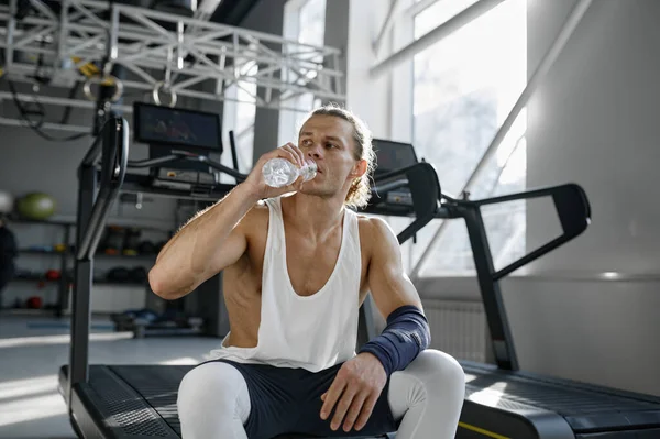 stock image Happy smiling athletic man drinking water taking break after workout in gym. Excited sportsman relaxing while sitting on treadmill machine at fitness center