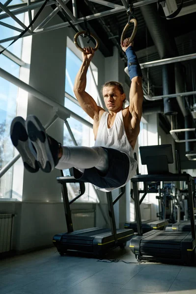 体育館のライトスポーツホールで体操リングに男性アスリートのトレーニングをフィット 強い腕を持つ筋肉の男は スポーツ機器にぶら下がっている間に足を持ち上げる運動を行う — ストック写真