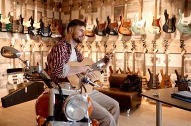 Müzik dükkanında motosiklette oturan mutlu, gülümseyen, akustik gitar çalan adam. Genç pazarlamacı müşteri çekiyor.