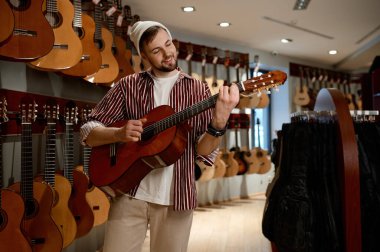 Genç erkek müşteri dükkanda akustik gitar seçiyor. Hippi adam enstrümanın boynuna dokunuyor. Cihazların yanında duruyor.