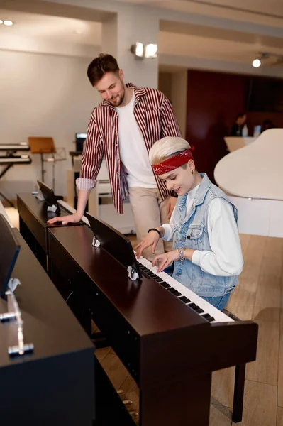 プロの店で1つを選択しながら楽器で演奏する若い女性ピアニスト セールスマンは近くの女性顧客に立つ選択を助けることを試みる — ストック写真