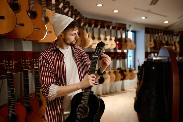 店でアコースティックギターを選ぶ若い男性客 装置の近くに立っている楽器の首に触れるヒップスター男 — ストック写真