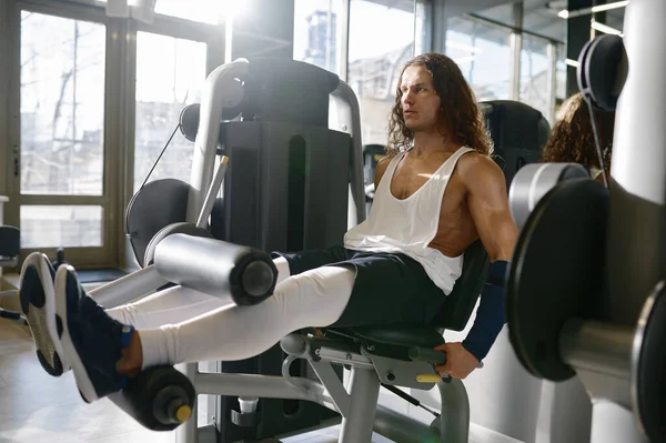 若い運動スポーツマンは ジムマシン上で筋肉を行使し 曲げ足をポンピング 背景にスポーツクラブのインテリア — ストック写真