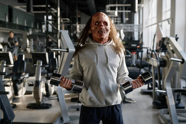 在现代体育俱乐部里 一个长着令人毛骨悚然的脸的僵尸男子用哑铃训练肌肉 — 图库照片