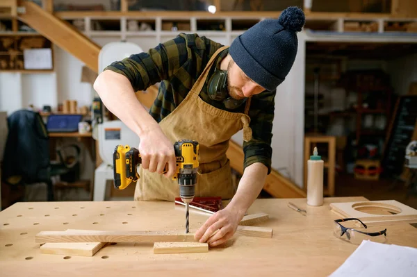 在木工车间里 与钻头一起工作的年轻人或工匠靠着桌子干活 用专业仪器组装木制零件的修理工 — 图库照片