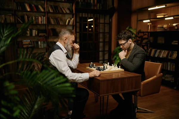 Senior man and young chess player having friendly match at home library. Enthusiastic old father playing with younger son enjoying logic board game